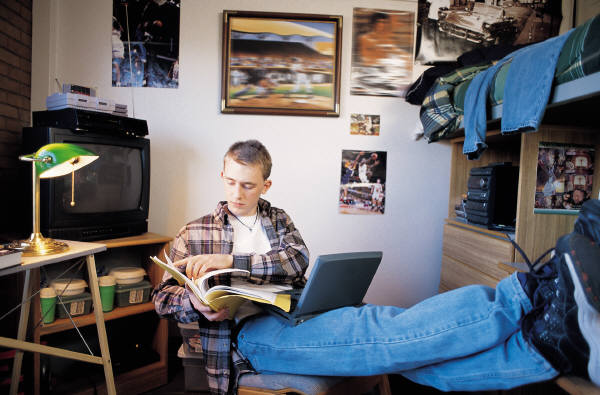 Picture of a male college student in his dorm room reading, with his feet up on the dresser